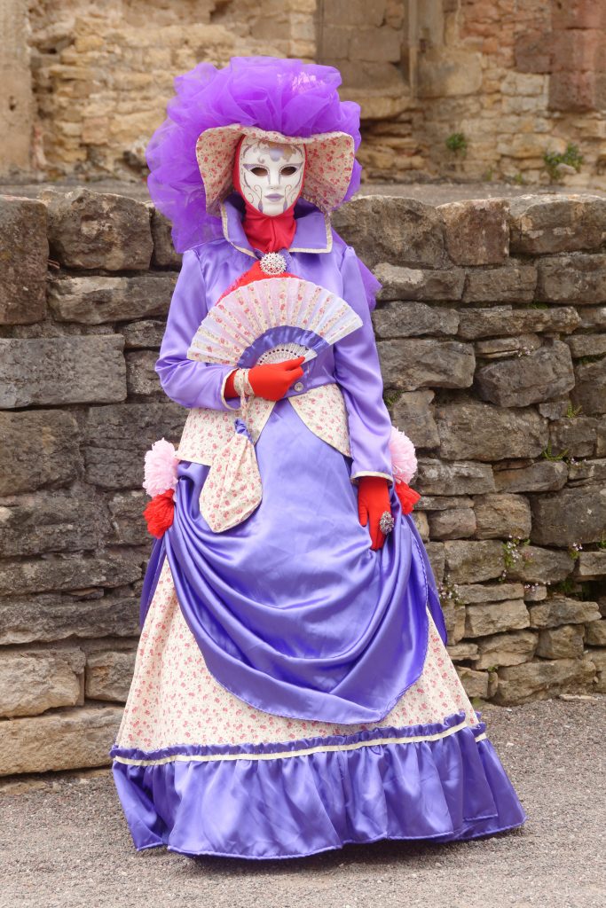 Costume vénitien violet à masque blanc et éventail, devant un mur de pierres