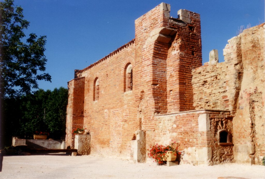 Ancien édifice en brique rouge aux murs partiellement ruinés, cour gravillonnée et géraniums fleuris