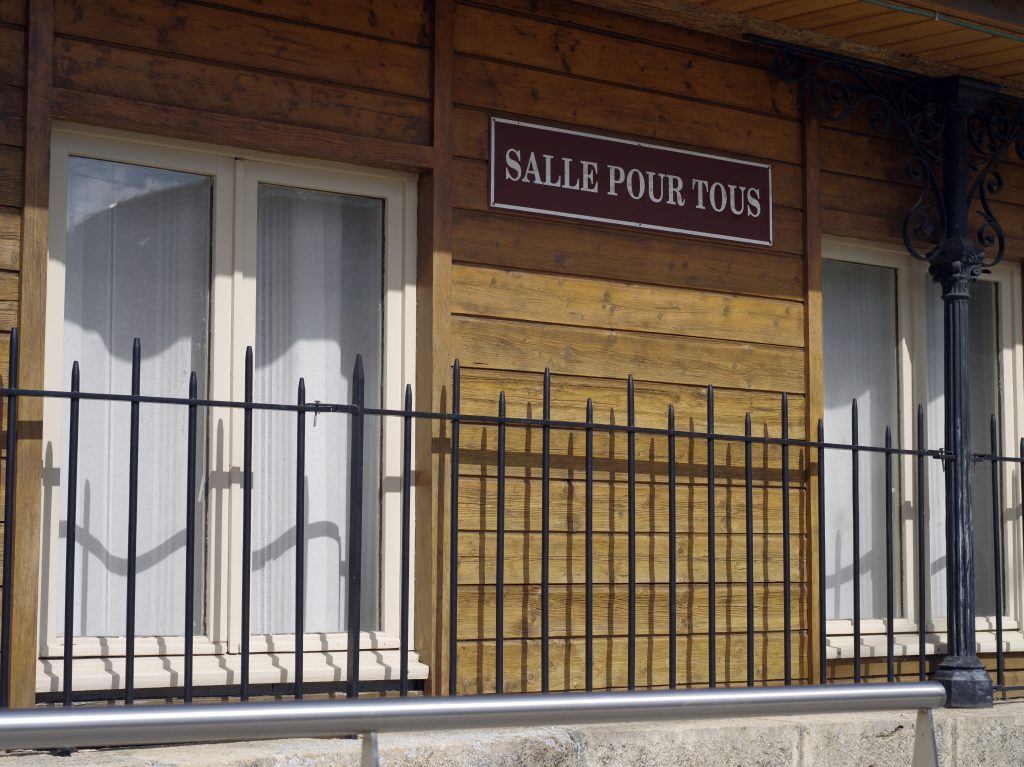 Plaque « Salle pour tous » sur une façade en bois derrière une grille noire