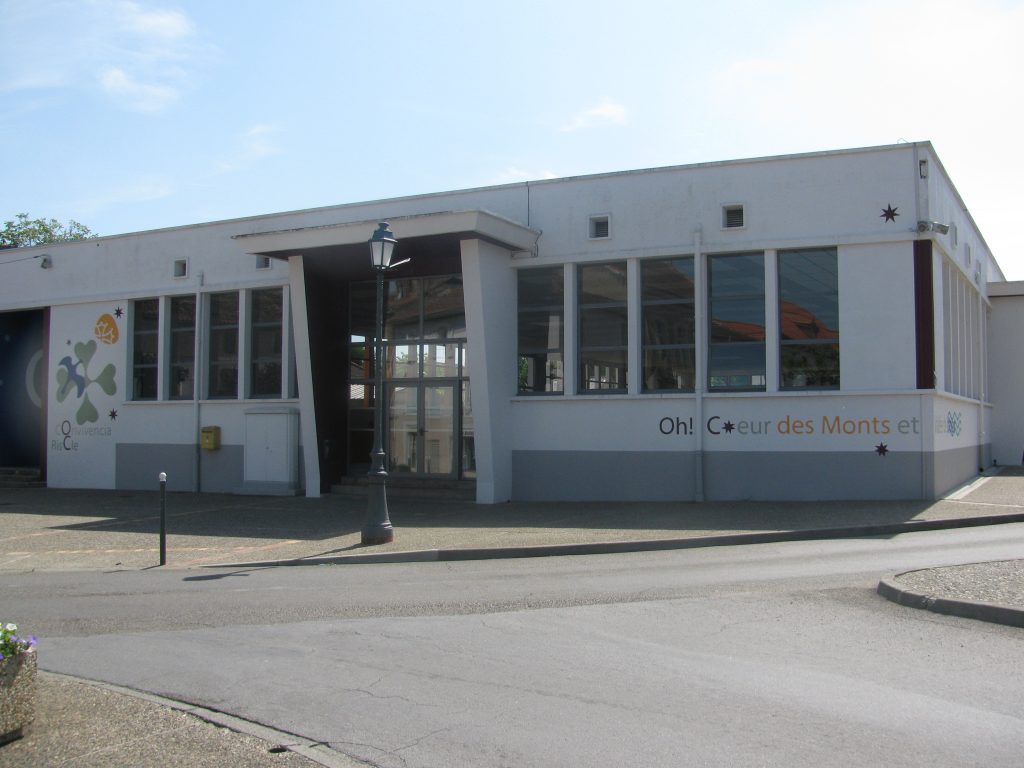 Bâtiment communal blanc aux larges baies vitrées, lampadaire et parking goudronné devant l'entrée