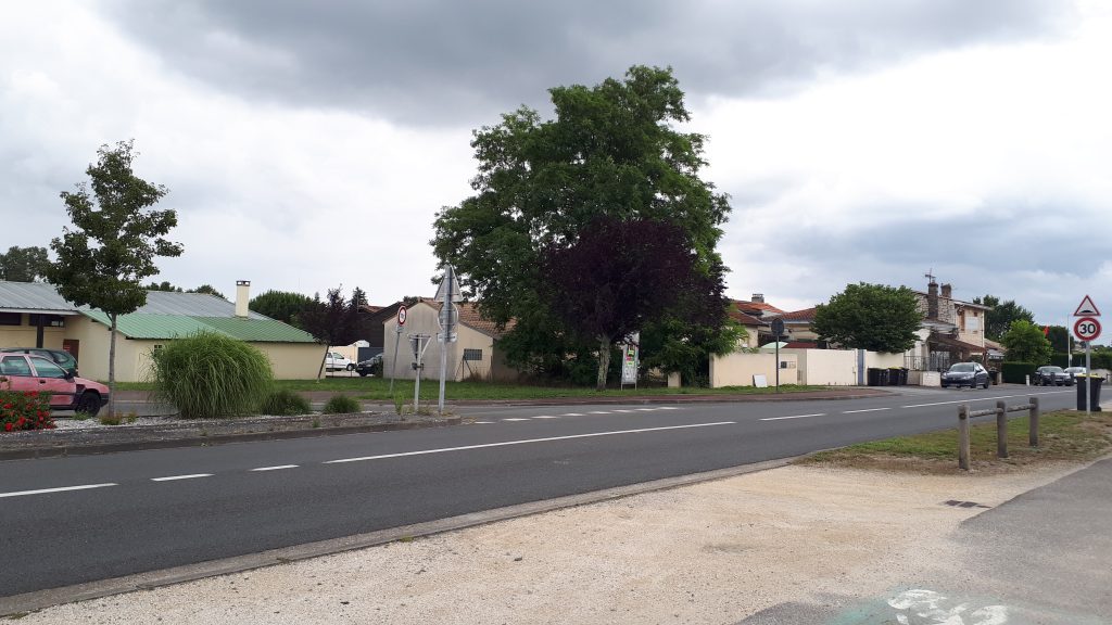 Route traversant un village, maisons basses et grands arbres sous un ciel nuageux, panneau 30