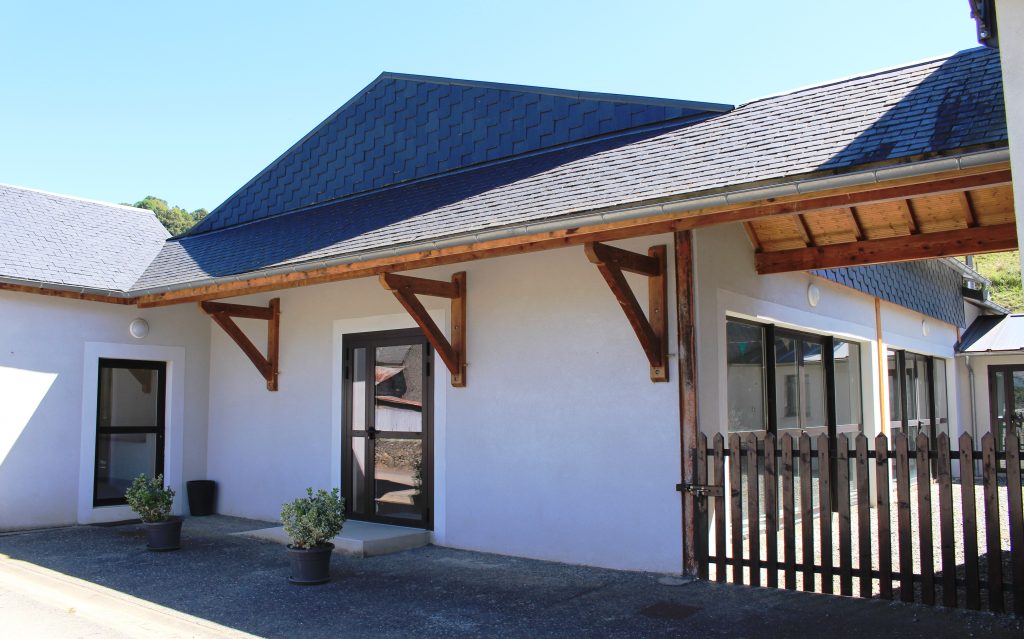 Bâtiment communal blanc à toit d'ardoises, auvent en bois, portes vitrées et arbustes en pot