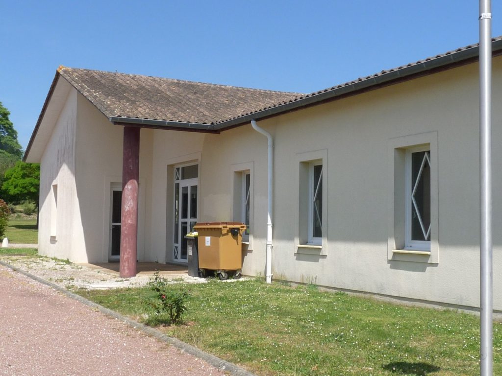 Salle communale beige à toit de tuiles, pelouse et conteneur à déchets devant l'entrée