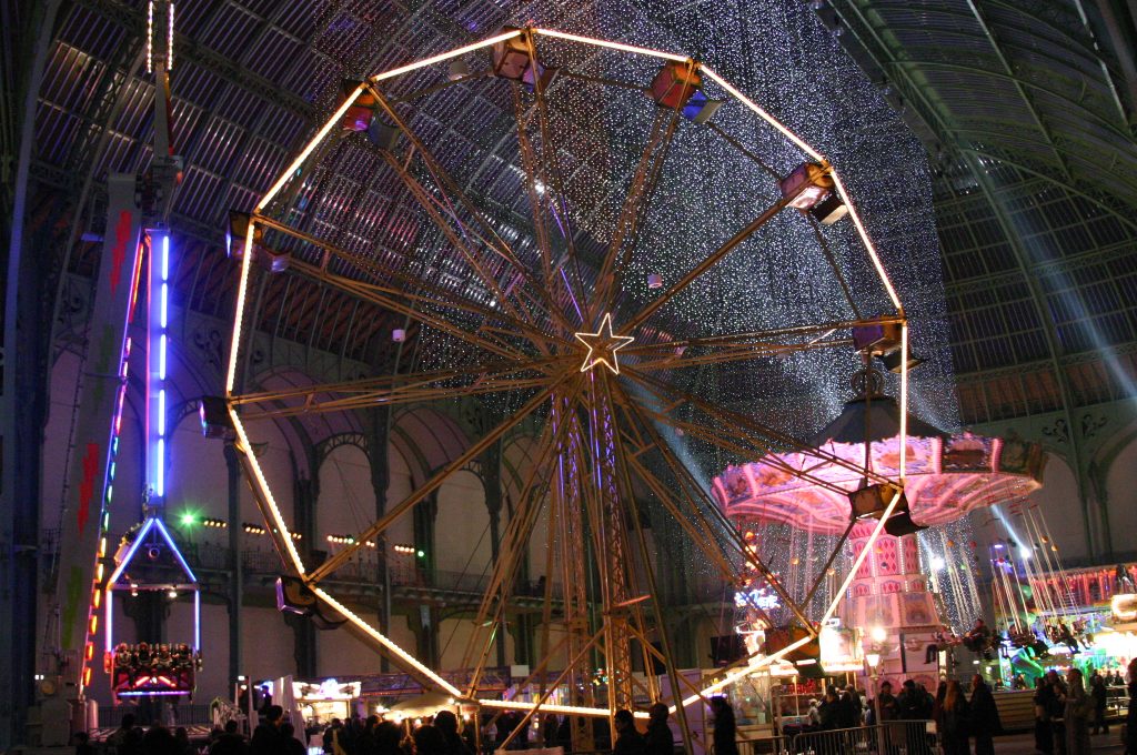 Grande roue illuminée et manège dans une halle vitrée, foule de visiteurs le soir