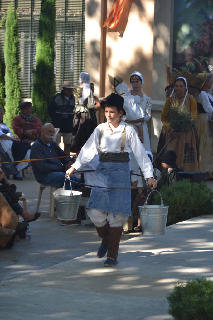 Jeune garçon en costume provençal portant deux seaux en zinc devant un public costumé