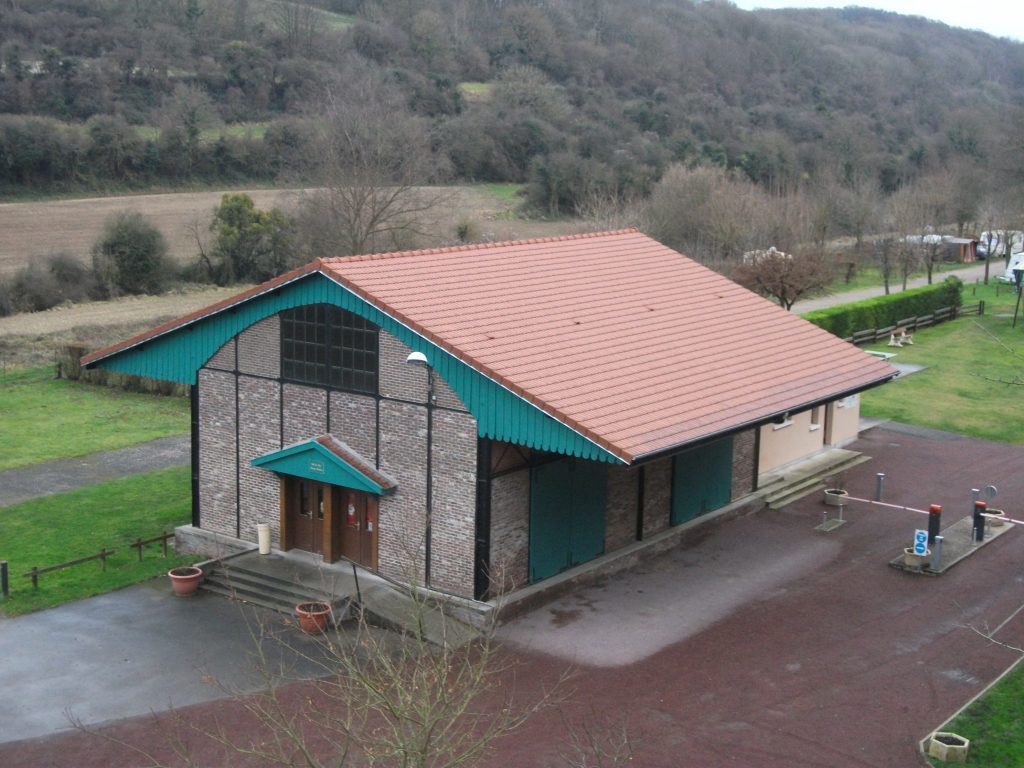 Salle des fêtes en brique au toit de tuiles rouges, bordée de champs et collines boisées
