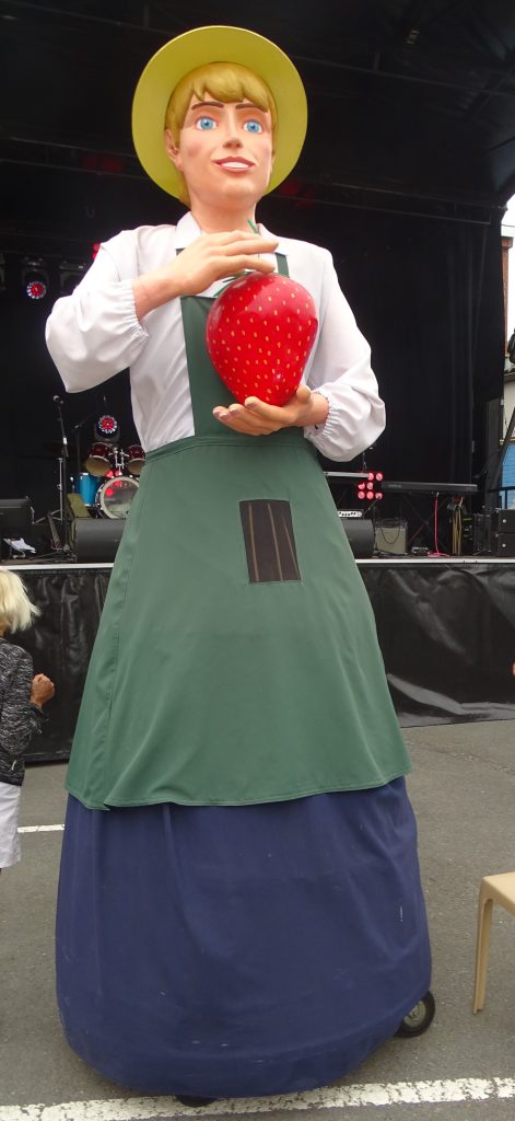Géant de procession en tablier vert tenant une grosse fraise devant une scène de concert