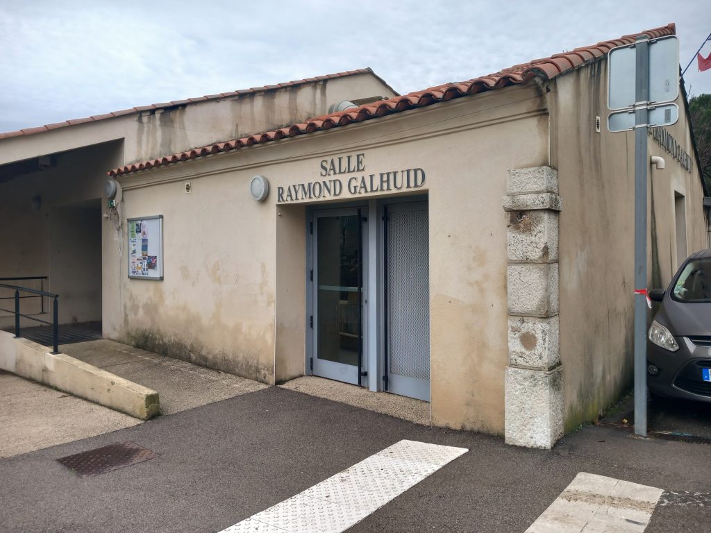 Façade de la salle Raymond Galhuid, bâtiment communal beige à toit de tuiles, voiture stationnée