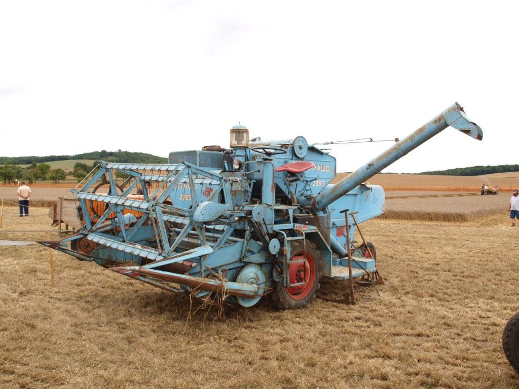 Ancienne moissonneuse-batteuse bleue exposée dans un champ moissonné, spectateurs au loin