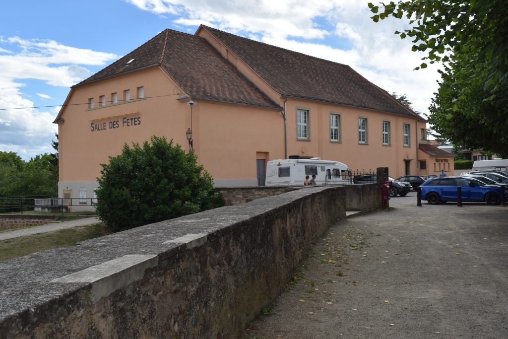 Salle des fêtes ocre au toit de tuiles, parking, camping-car et muret de pierre