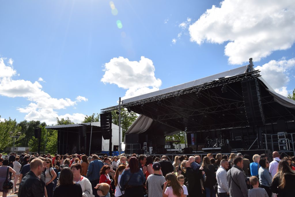Foule rassemblée devant une grande scène de concert en plein air, ciel bleu nuageux