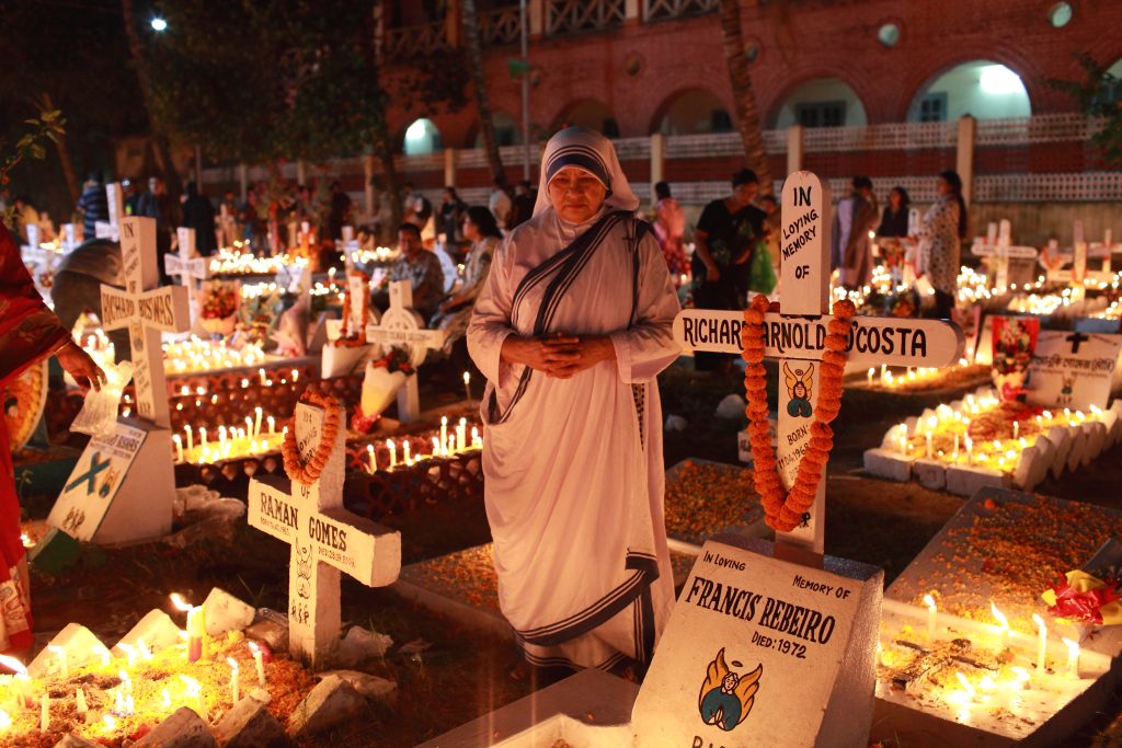 Religieuse en sari blanc recueillie parmi des tombes fleuries illuminées de bougies, la nuit