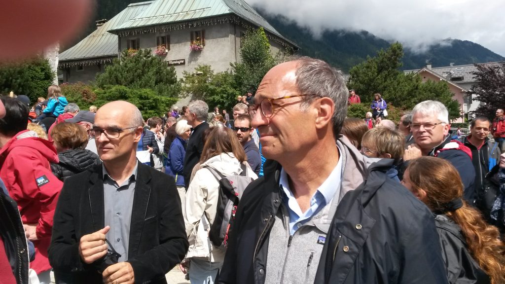 Foule en plein air devant un chalet fleuri, montagnes boisées et nuages en arrière-plan