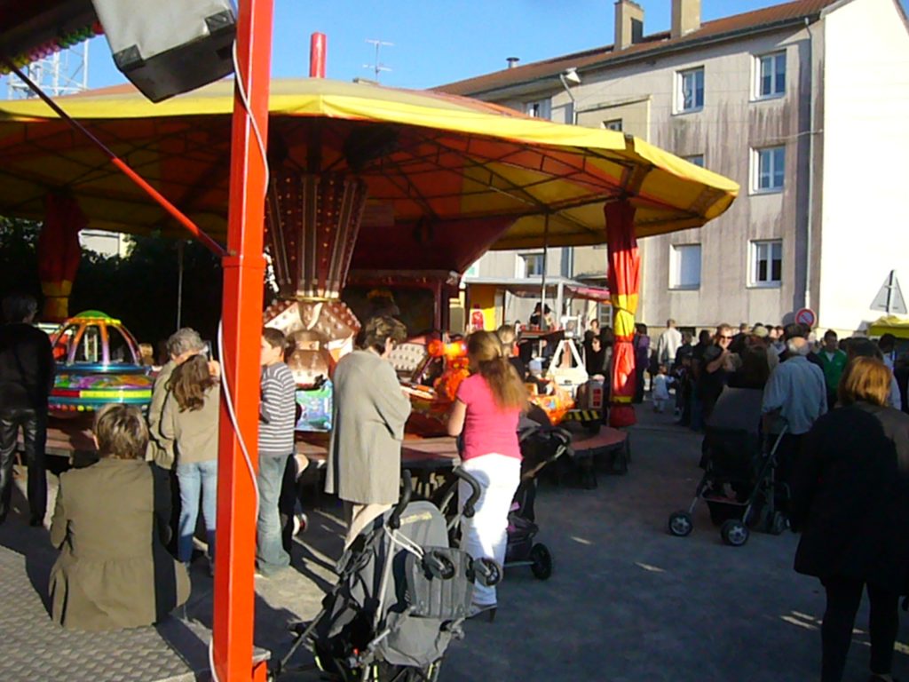 Manège forain jaune et rouge entouré de familles et poussettes sur une place