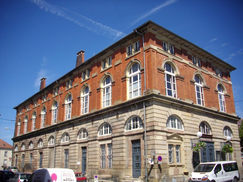 Grand bâtiment en brique rouge et pierre, salle des fêtes, ciel bleu, véhicules garés