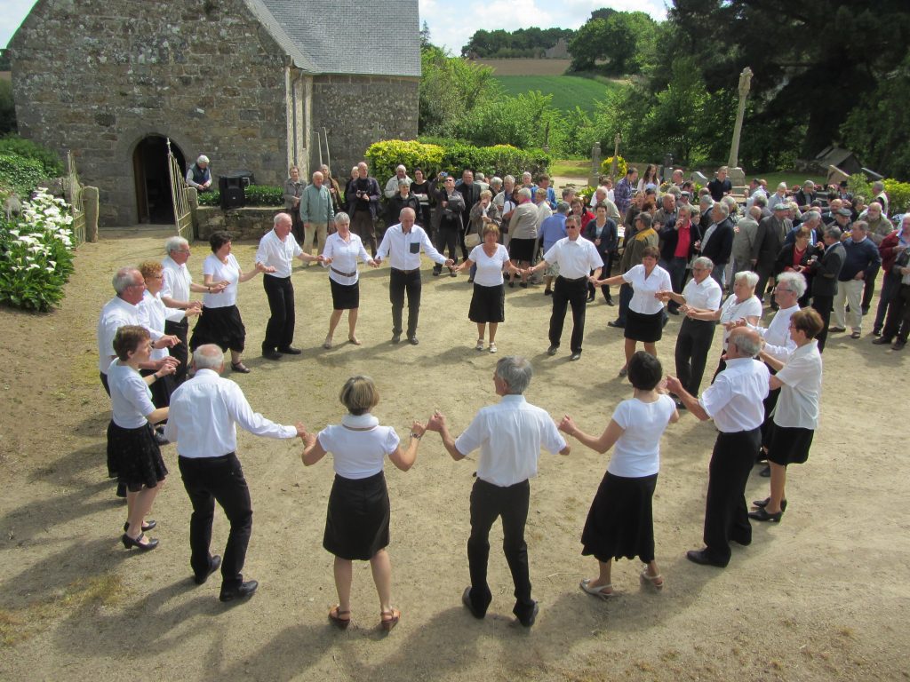 Danseurs en ronde devant une chapelle en pierre, public rassemblé autour