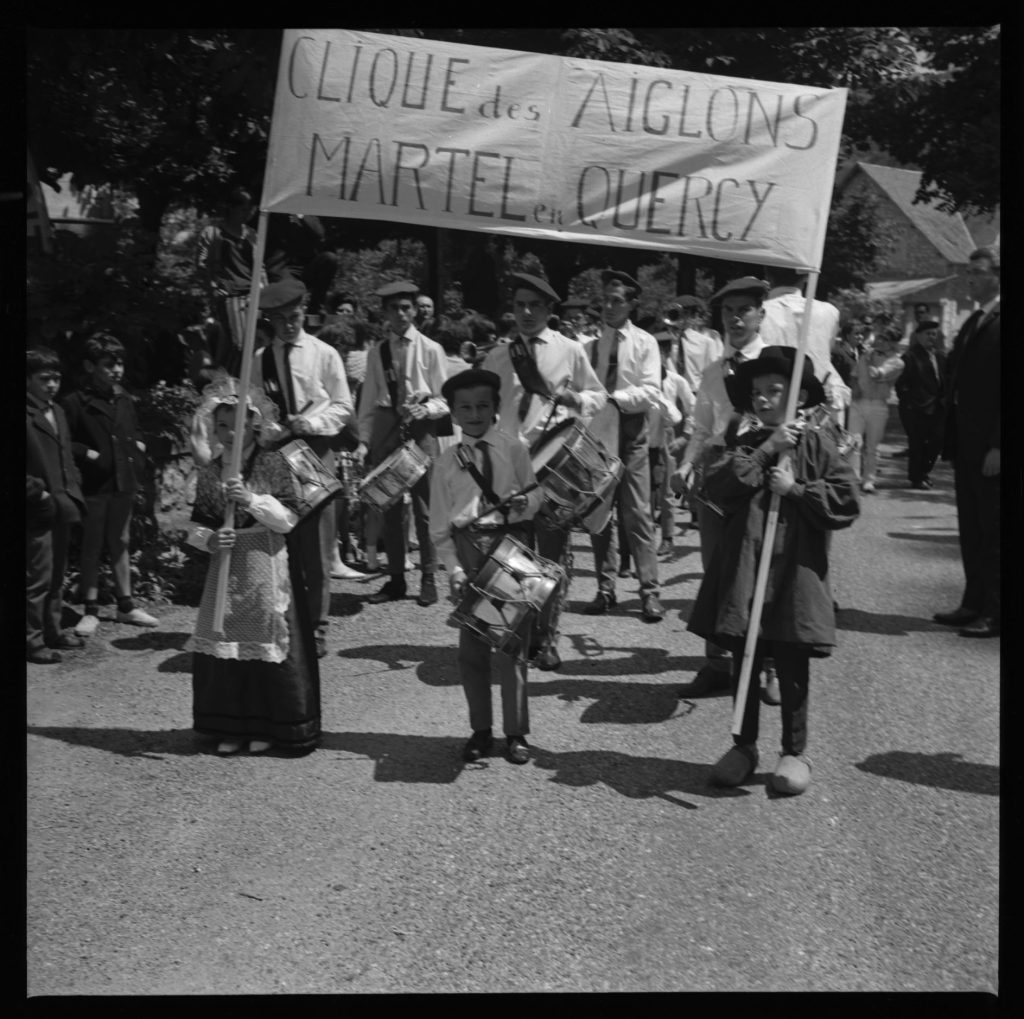 Défilé en noir et blanc d'une clique de tambours derrière une banderole, enfants en costume