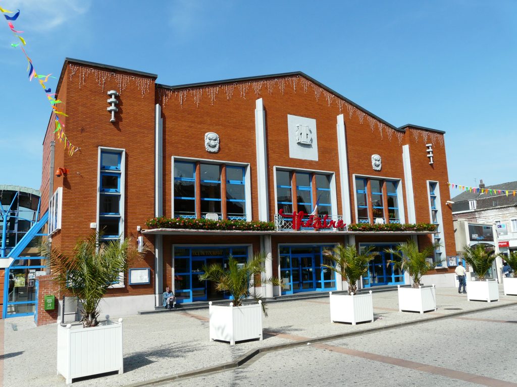 Théâtre en brique rouge aux menuiseries bleues, palmiers en bacs et guirlandes sur la place