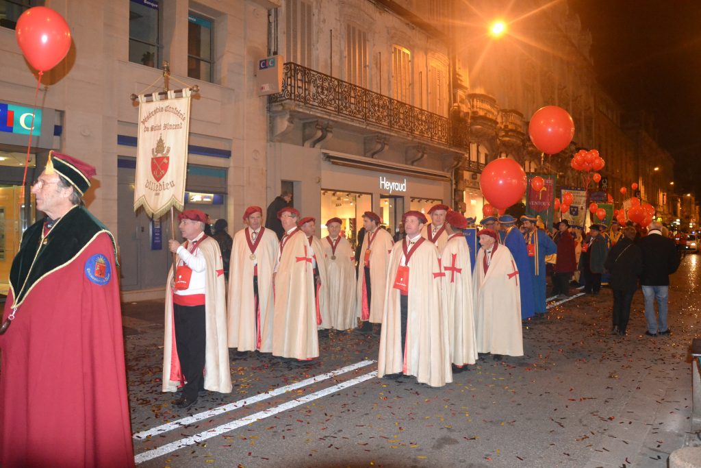 Confrérie en capes crème et coiffes rouges défilant avec bannière et ballons, rue nocturne