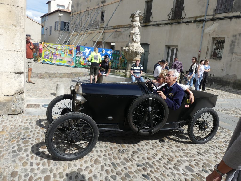 Voiture ancienne noire décapotable avec deux occupants sur une place pavée de village