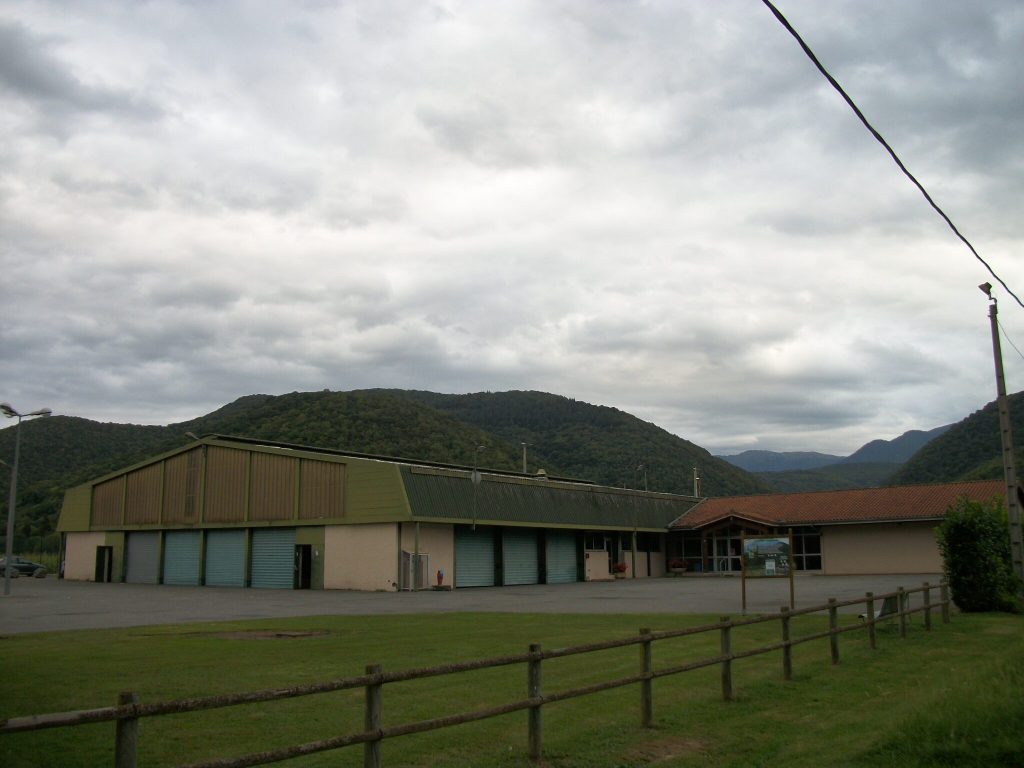 Grand hangar communal et sa salle d'accueil au pied de collines boisées, ciel nuageux