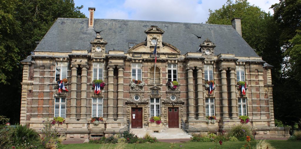 Mairie en pierre et brique pavoisée de drapeaux français, balconnières fleuries, pelouse arborée